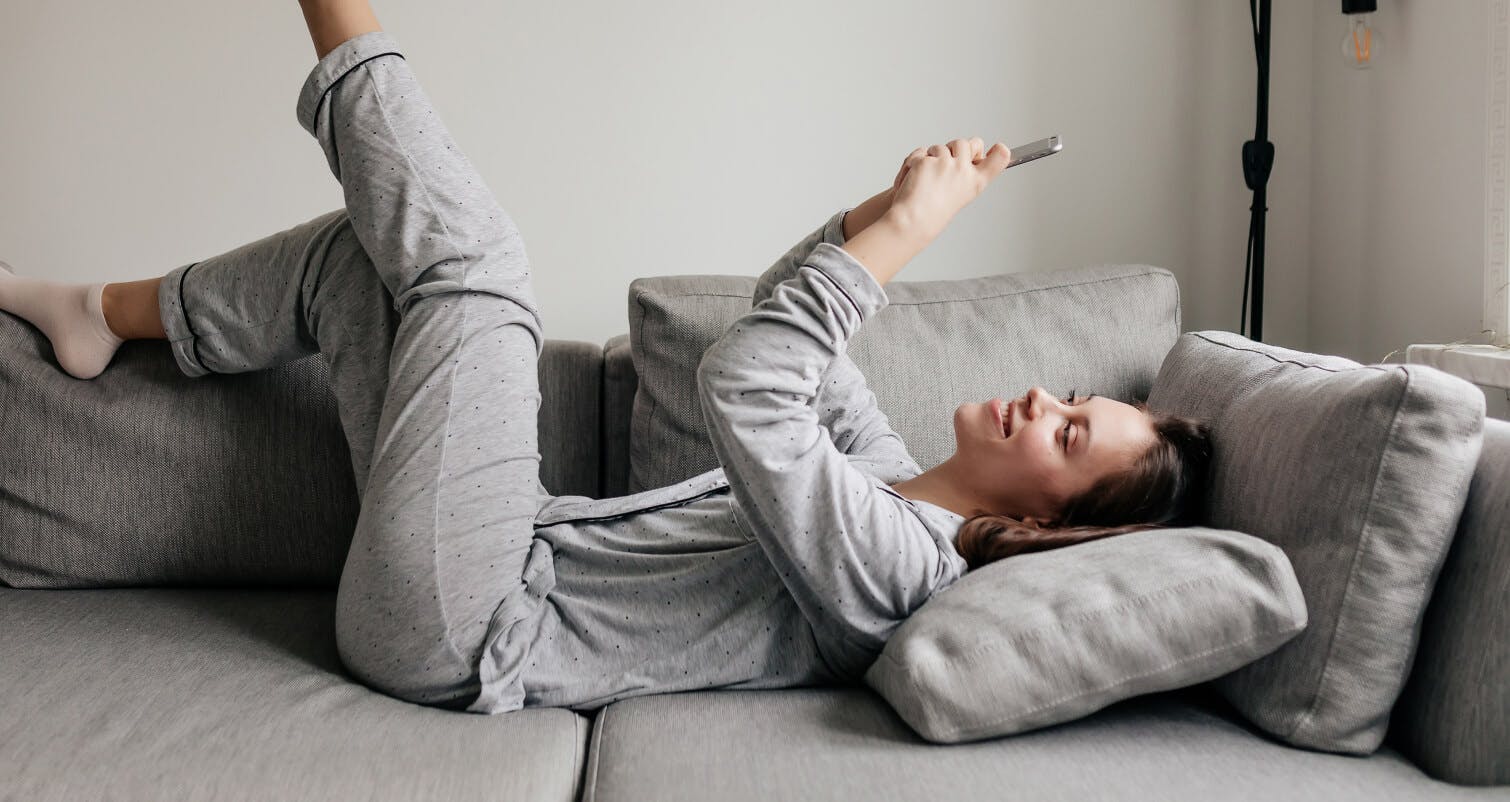 Mulher de pijama deitada em um sofá cinza digitando no celular